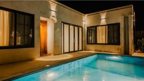 A swimming pool with a lighted deck area, a door, and windows with curtains.