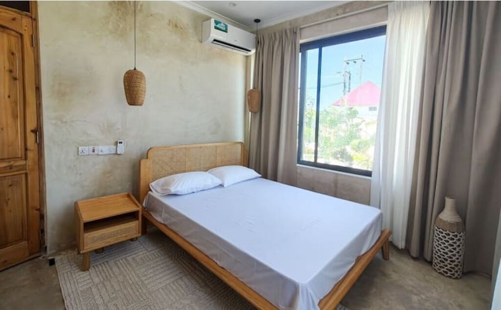 Un dormitorio con una cama individual, una mesita de noche de madera, una lámpara de fibras naturales, un equipo de aire acondicionado y una ventana con cortinas.