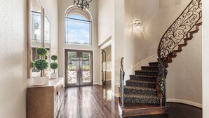 A spacious hallway with a wooden floor, a grand staircase with ornate railings, and a large arched window allowing natural light. - Waterfront home, 6 bedrooms private pool and dock on Lake Conroe (Montgomery)