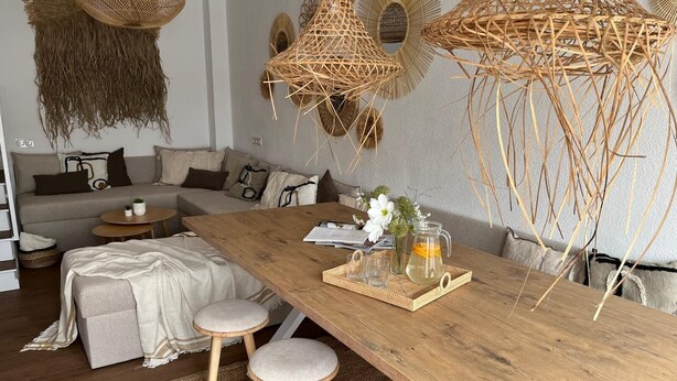 Een moderne woonkamer met een houten eettafel, twee krukken, een bank en decoratieve wandkleden.
