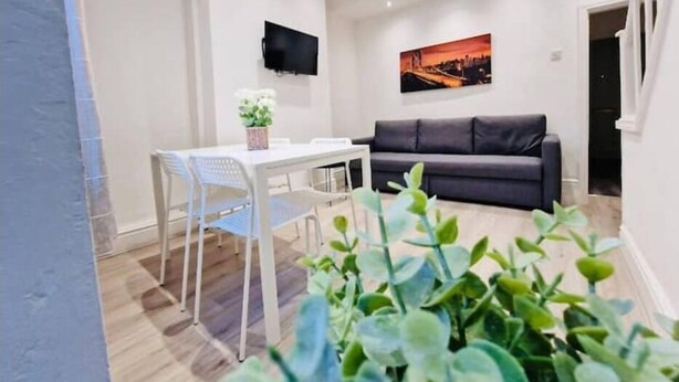 Una sala de estar moderna con una mesa de comedor blanca, un sofá negro y un televisor de pantalla plana montado en la pared.