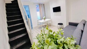 Una escalera conduce a un área de comedor con una mesa y sillas blancas, un sofá gris y un taburete negro pequeño.