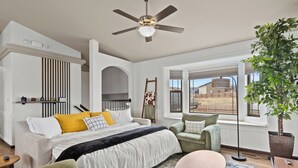 A bedroom with a bed, a chair, a table, a floor lamp, and a large window with a view of the outside.