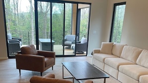 A spacious living room with a large sliding glass door leading to an outdoor deck, a brown leather sofa, a white sofa, and a coffee table. - Brand NEW MINT Windham 5BR/4.5BA home on 5 acre, wooded hilltop. (Windham)