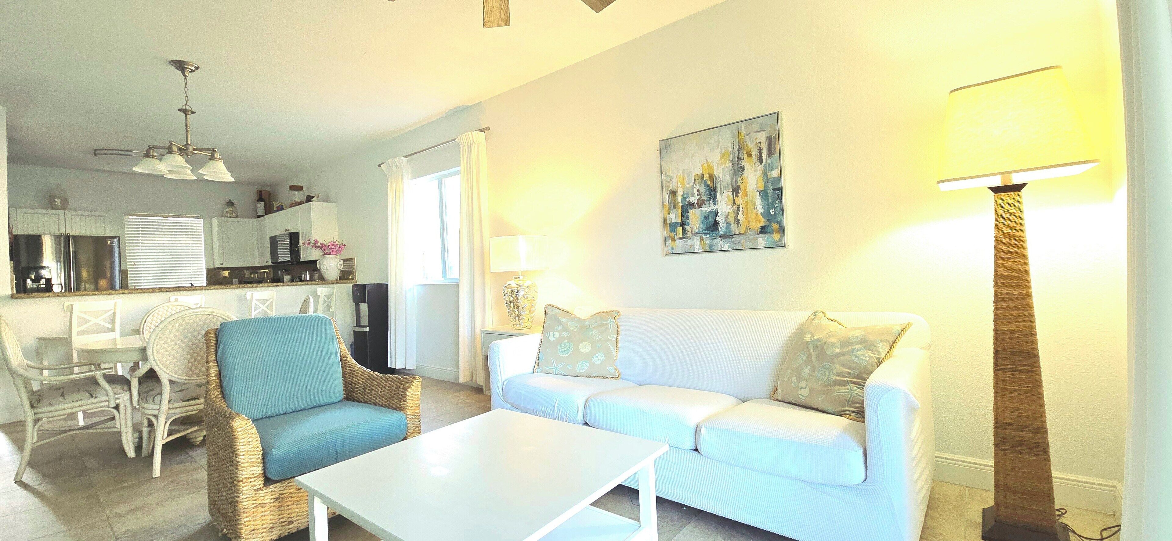 A living room with a white sofa, blue armchair, and a coffee table. There is a dining area with a table and chairs, and a kitchen in the background.
