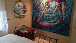 Un dormitorio con cama, espejo redondo, ventilador y un cuadro abstracto grande en la pared.