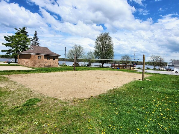 Una cancha de voleibol de arena con un edificio de piedra, una cerca de madera y un lago al fondo.