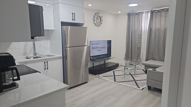 A modern kitchen with a stainless steel refrigerator, a built-in oven, and a microwave. There's a flat-screen TV mounted on the wall displaying news. The room has a light-colored floor and a patterned rug. - Rome Sweet Home 2. Private Guest Suite in a quiet, family-friendly neighborhood. (Calgary)