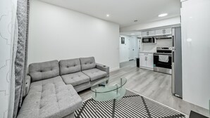 A modern living room with a grey sofa, a glass coffee table, and a kitchen area with white cabinets and appliances. - Rome Sweet Home 2. Private Guest Suite in a quiet, family-friendly neighborhood. (Calgary)