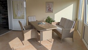 Una habitación iluminada por el sol con una mesa de vidrio rodeada de sillas de mimbre, una planta en maceta y un cuadro en la pared.