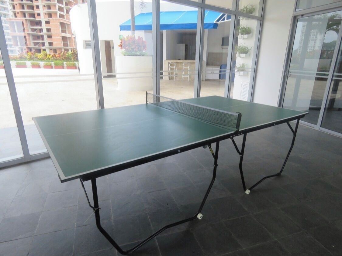 Taula de ping-pong en un espai interior modern amb grans finestres, vista d'un edifici a l'exterior i una tenda blava visible a través de les finestres.