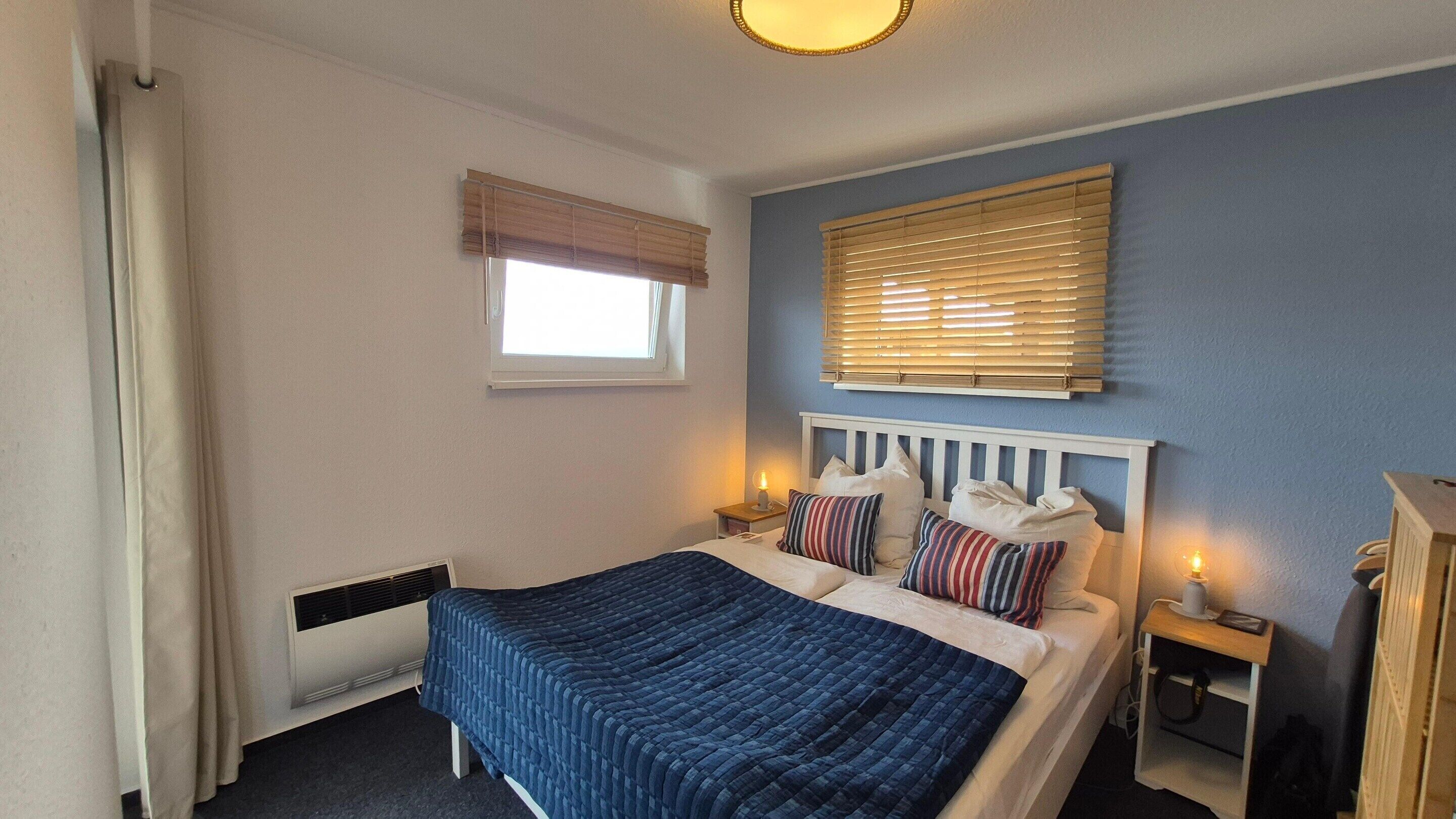 A bedroom with a bed, bedside tables, a window with blinds, a radiator, and a wall-mounted light fixture.
