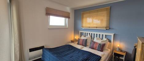 A bedroom with a bed, bedside tables, a window with blinds, a radiator, and a wall-mounted light fixture.