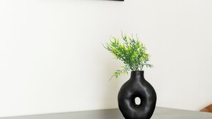 A table with a black vase containing green plants, wooden chairs, and a framed painting on the wall. - Elysee apartment 7 (Athens)