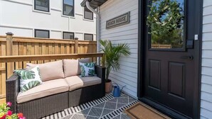 A front view of a house with a 'BEACH HOUSE' sign, a black door, a wooden fence, a wicker sofa with cushions, a potted plant, and a metal watering can. - Charming 3BR Beach House with BBQ in Grand Bend (Grand Bend)