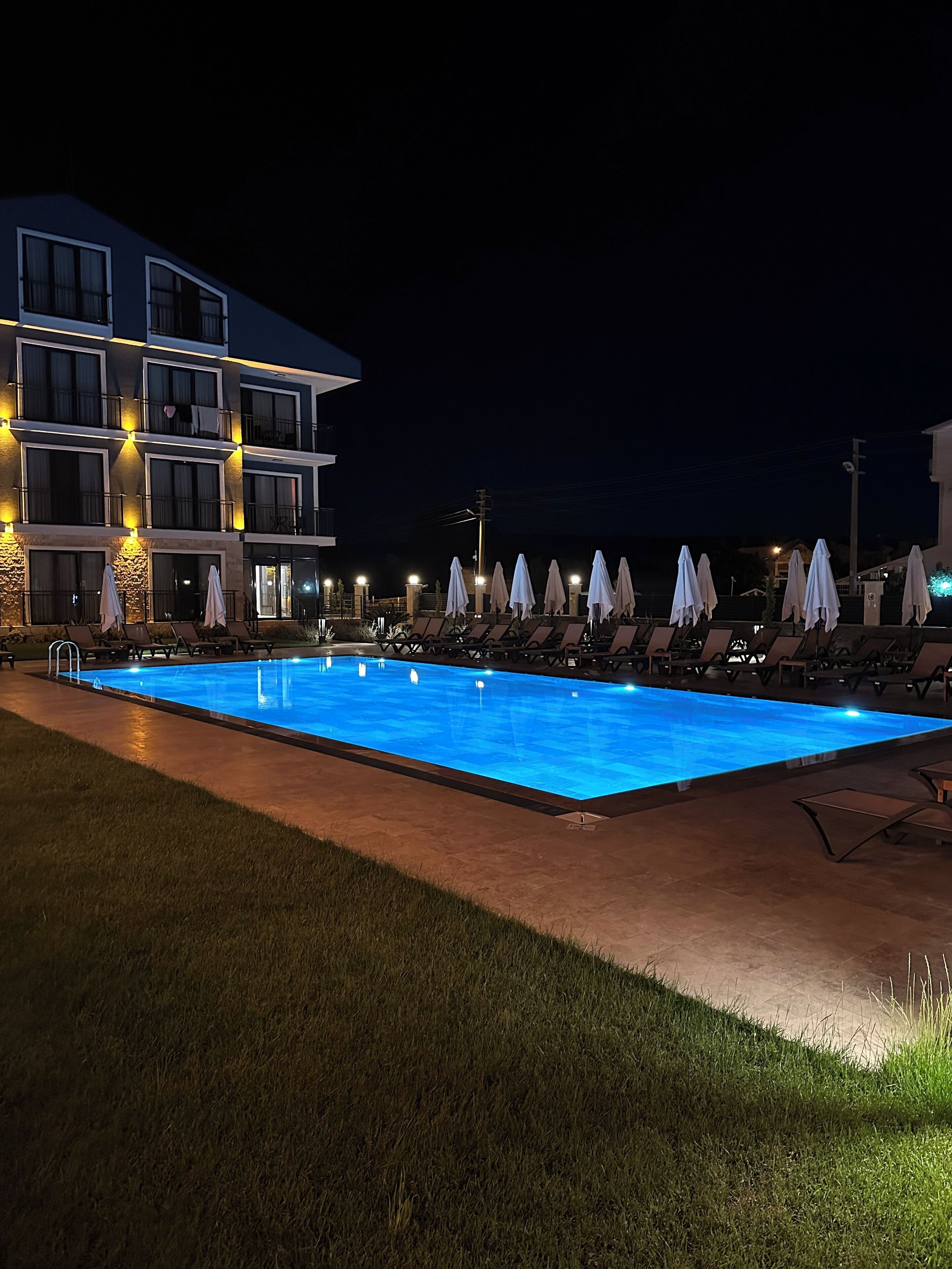 A nighttime swimming pool area with lounge chairs and umbrellas, surrounded by a building with multiple balconies and lights.