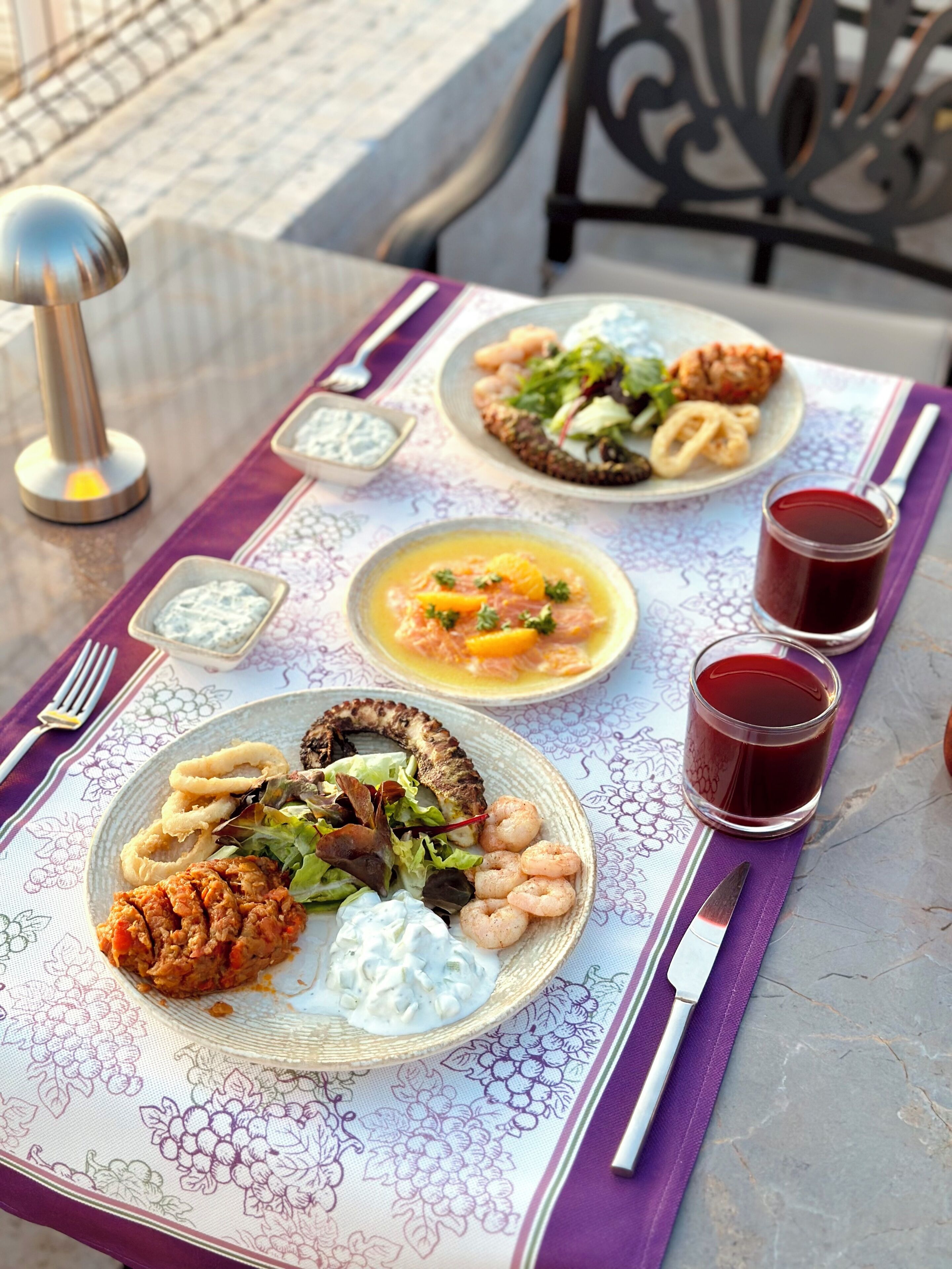 A table set with plates of food, including seafood and salad, and two glasses of red beverage.