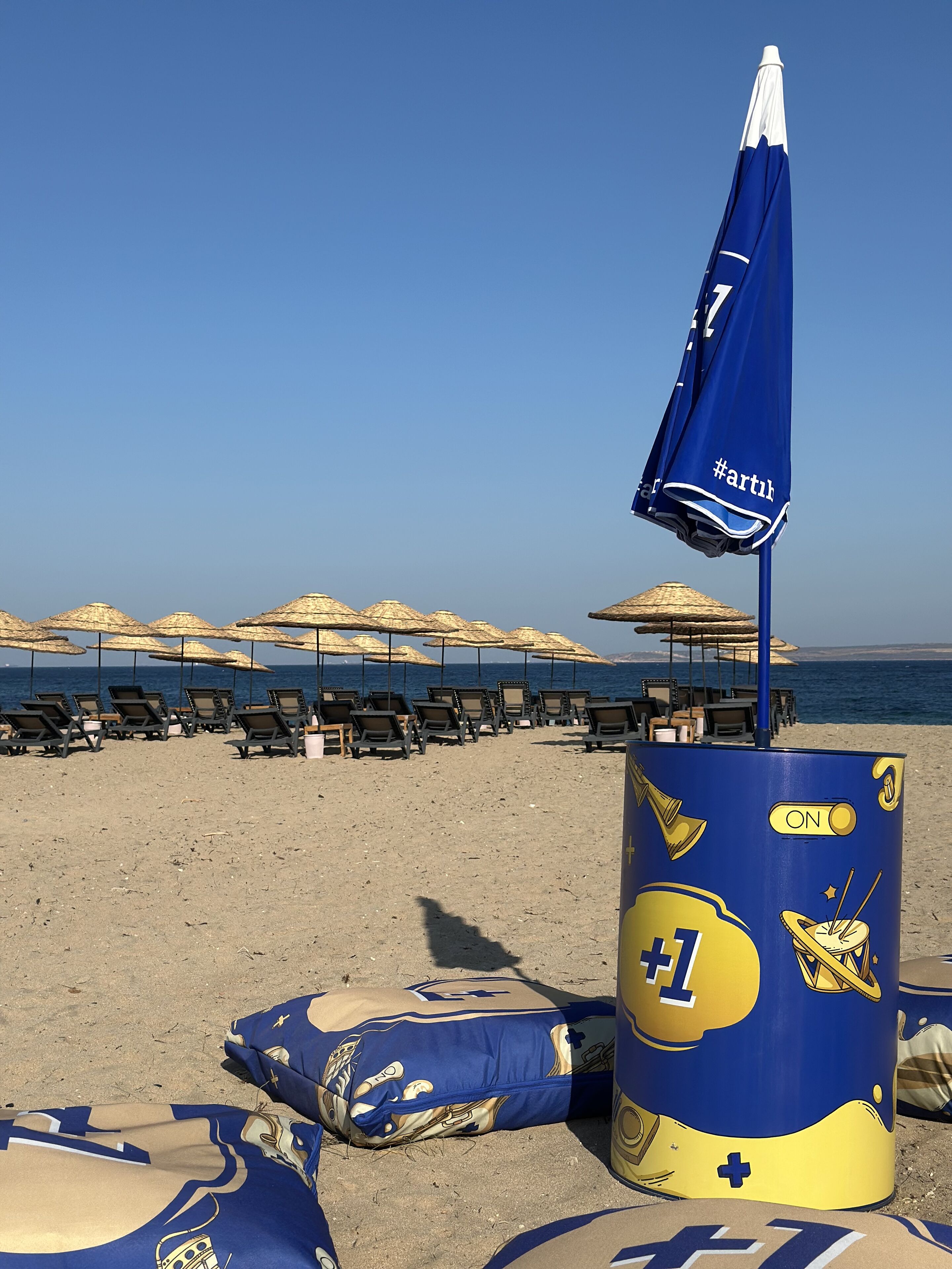 A beach with rows of sun loungers and thatched umbrellas, a blue and yellow barrel with a hashtag, and a clear blue sky.