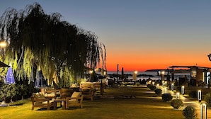 An outdoor seating area with wooden furniture and umbrellas, a large tree, and a sunset in the background. - GRAPE HOTEL (Ezine)