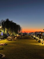 Un área de estar al aire libre con muebles de madera y sombrillas, un árbol grande y un atardecer de fondo.