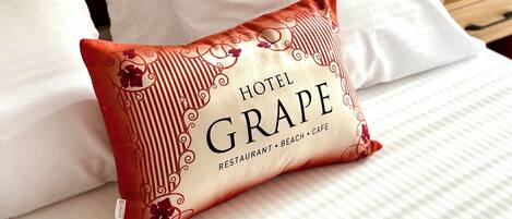 A hotel bed with white bedding and a decorative pillow labeled "Hotel Grape."