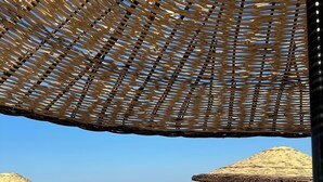 A beach with two thatched umbrellas, lounge chairs, and a small table. - GRAPE HOTEL (Ezine)