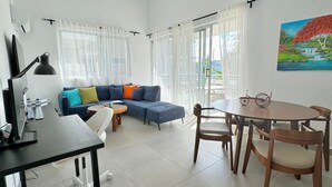 A modern living room with a blue sofa, a dining table with chairs, and a large window with sheer curtains.