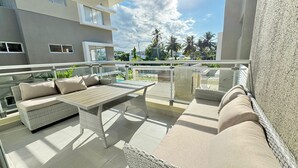 A balcony with wicker furniture, including a sofa and a table, overlooking a pool and palm trees.
