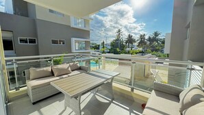 A balcony with wicker furniture, a glass railing, and a view of a pool and palm trees.
