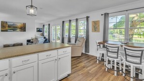 A kitchen with a wooden countertop, white cabinets, a dining area with a table and chairs, and large windows with curtains.