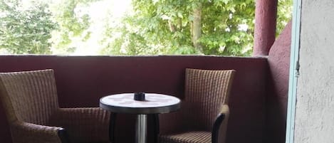 Un balcón con sillas de mimbre y una mesita redonda, con vistas a una zona verde con árboles.