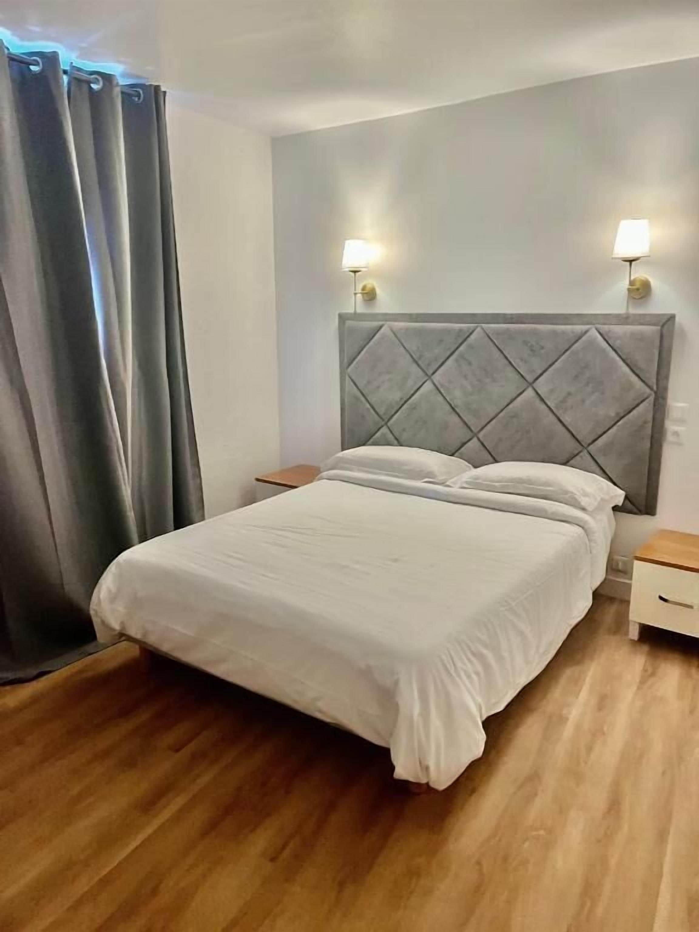 A bedroom with a bed, wooden floor, grey curtains, and two bedside lamps.