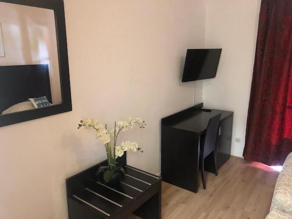 A hotel room with a bed, a desk with a flat-screen TV, a vase with flowers, and a mirror on the wall.