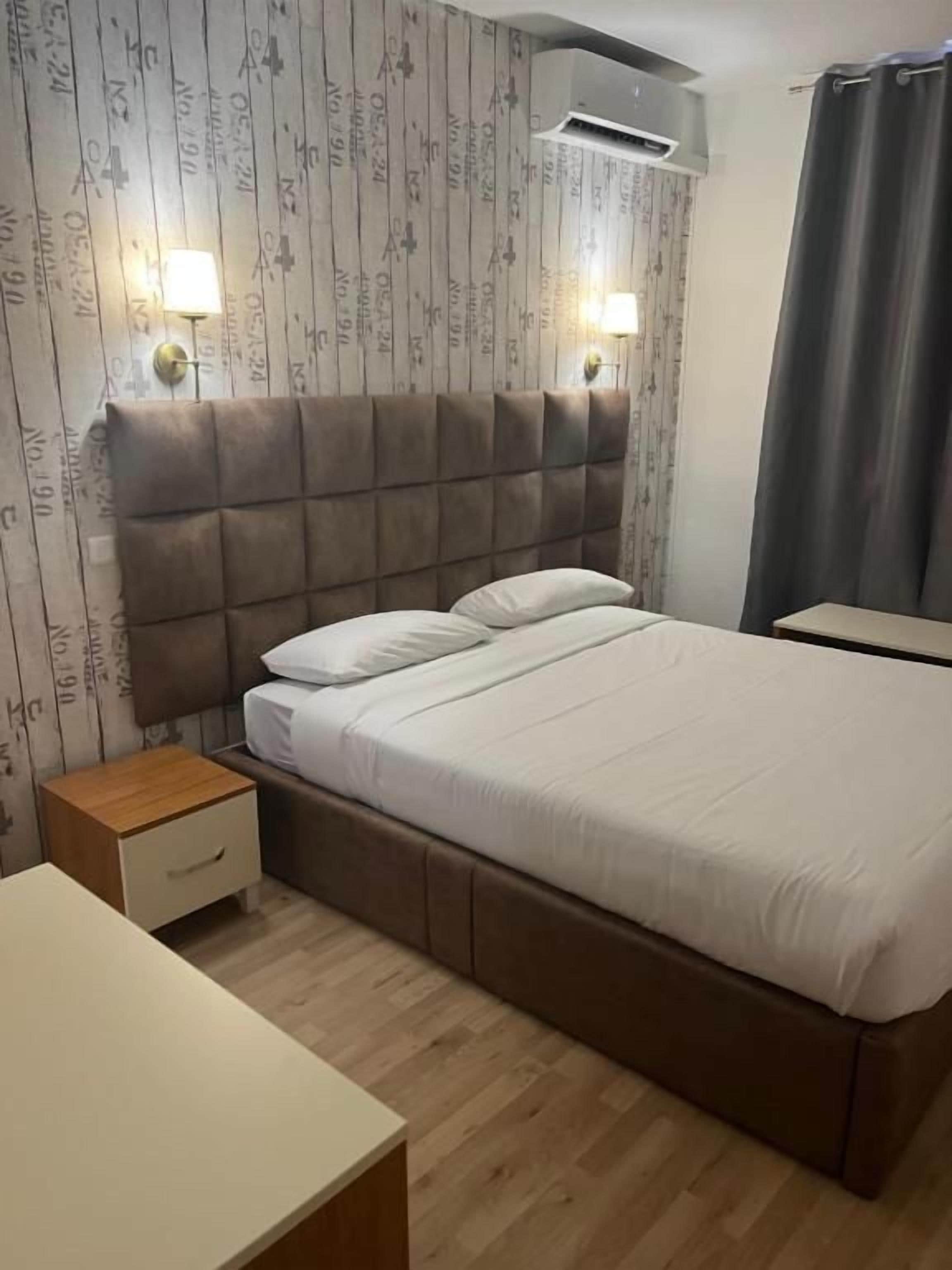 A bedroom with a bed, bedside tables, a nightstand, a wall-mounted air conditioner, and a curtain.