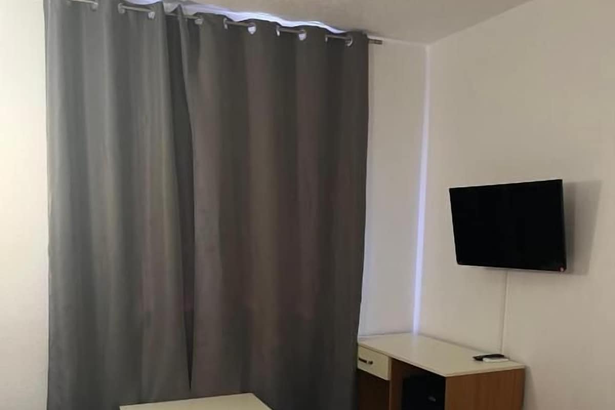 Una habitación de hotel con cama, escritorio, silla, televisor y cortina.