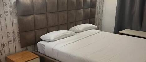 A bedroom with a bed, bedside tables, a nightstand, a wall-mounted air conditioner, and a curtain.