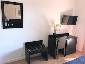 A room with a bed, a desk with a chair, a small table, a mirror, and a TV on the wall.