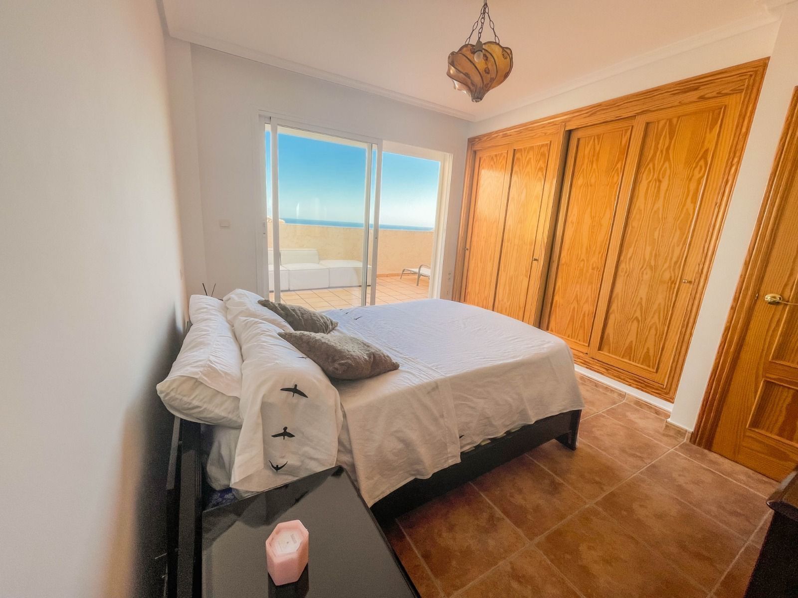 Une chambre à coucher avec un lit, une table de chevet sur laquelle est posée une bougie, une vue sur la mer par une porte coulissante et des armoires en bois.