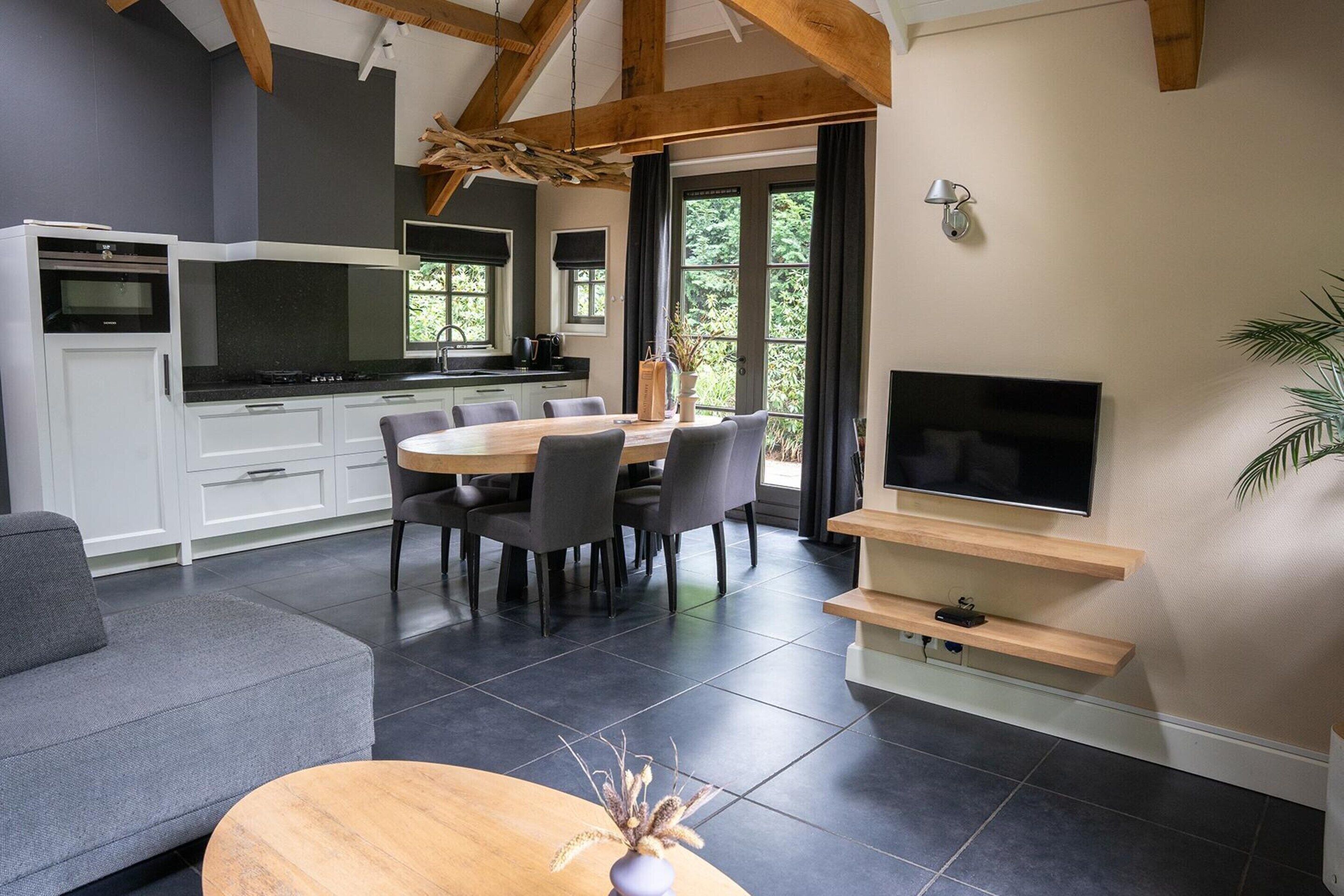 A modern kitchen with white cabinets, a dining area with a round table and chairs, a flat-screen TV mounted on a wooden shelf, and a grey sofa.