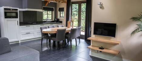 A modern kitchen with white cabinets, a dining area with a round table and chairs, a flat-screen TV mounted on a wooden shelf, and a grey sofa.
