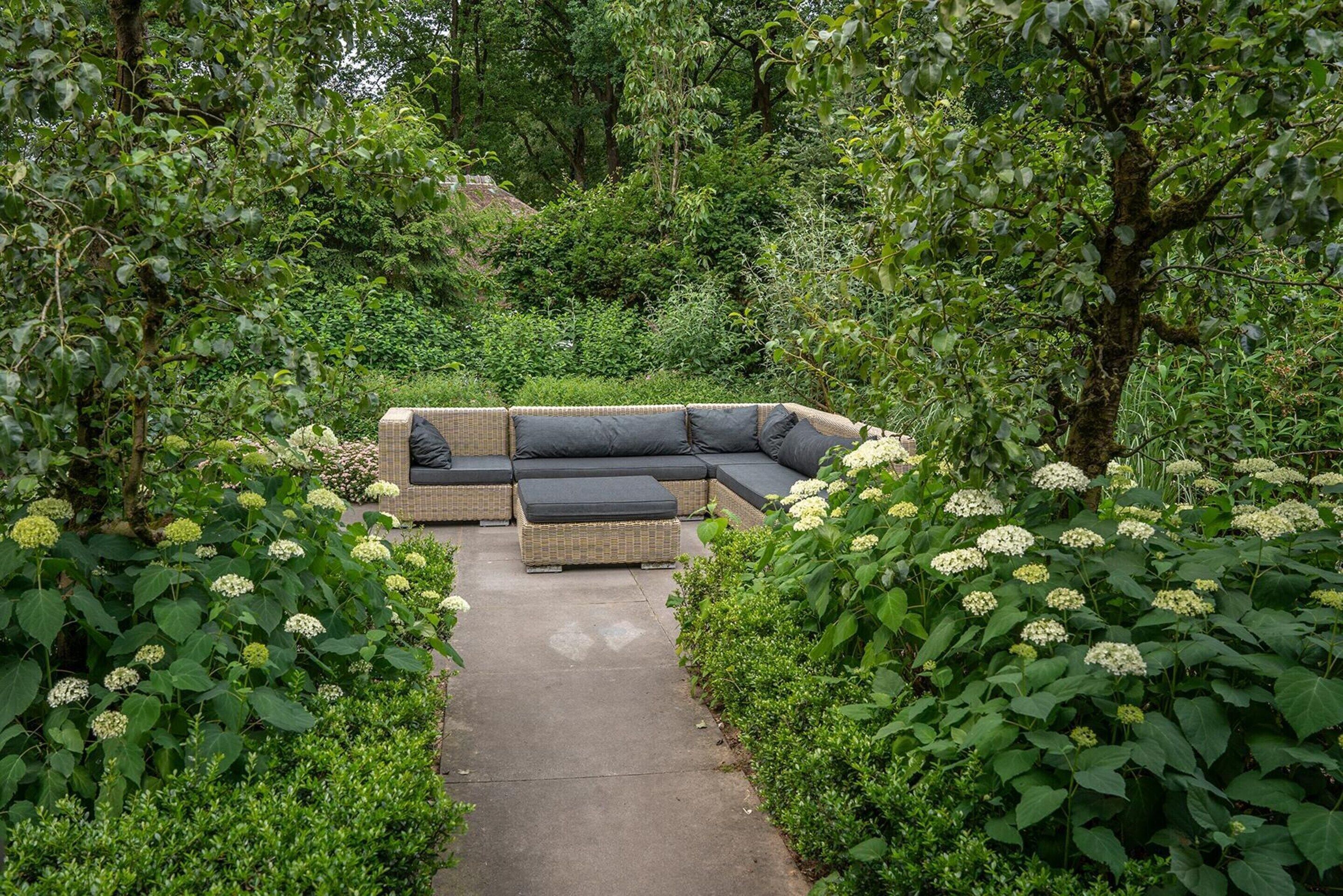 Ein Garten mit Ecksofa und Hocker, umgeben von üppigem Grün und weißen Blüten.