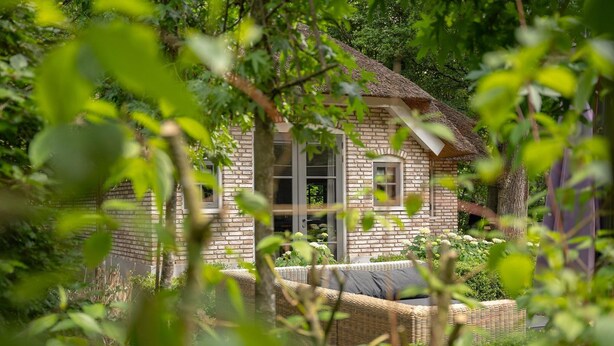 A rustic brick house with large windows, surrounded by lush greenery and a wicker sofa. - Farmhouse in Veluwe With Sauna and Nature Views (Ermelo)