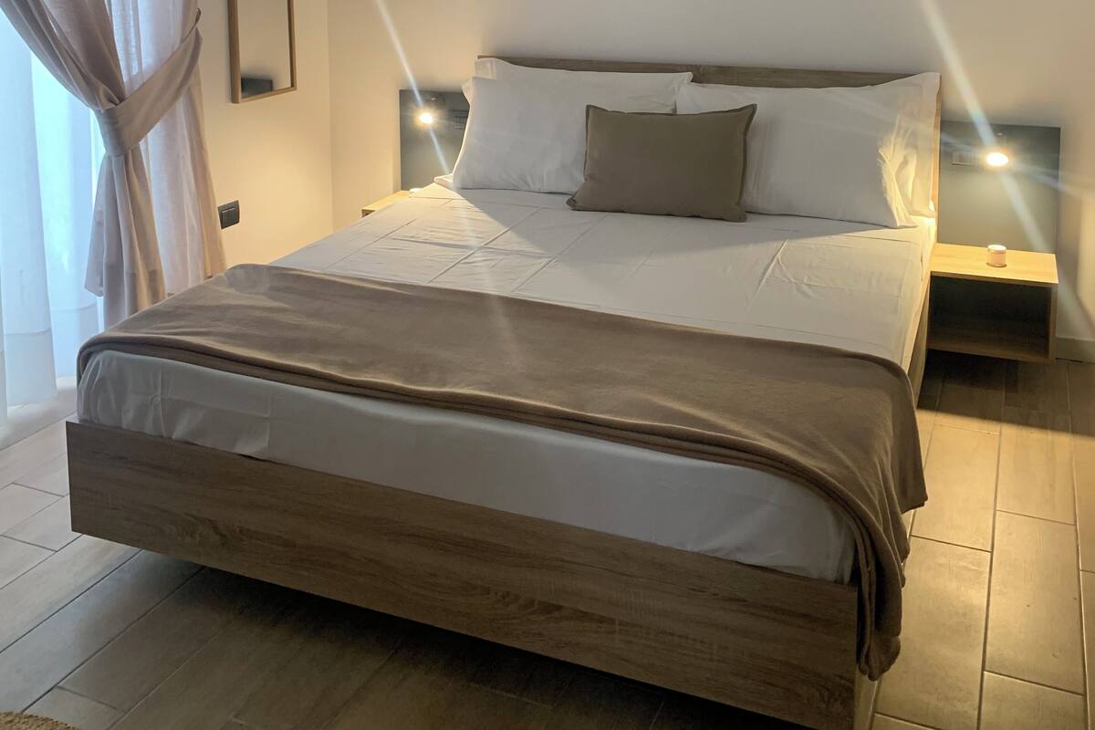 A modern bedroom with a large bed, a headboard, a bedside table with a lamp, and a wall-mounted artwork.