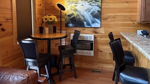 Interior acogedor de una cabaña con detalles de madera, una mesa redonda con sillas, un ventanal con persianas, un aire acondicionado de pared y un cuadro de una cascada.