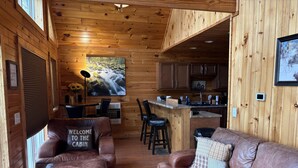 Interior de una cabaña con revestimiento de madera, un sofá, una mesita de centro, una zona de cocina y un ventanal con vistas a una cascada.
