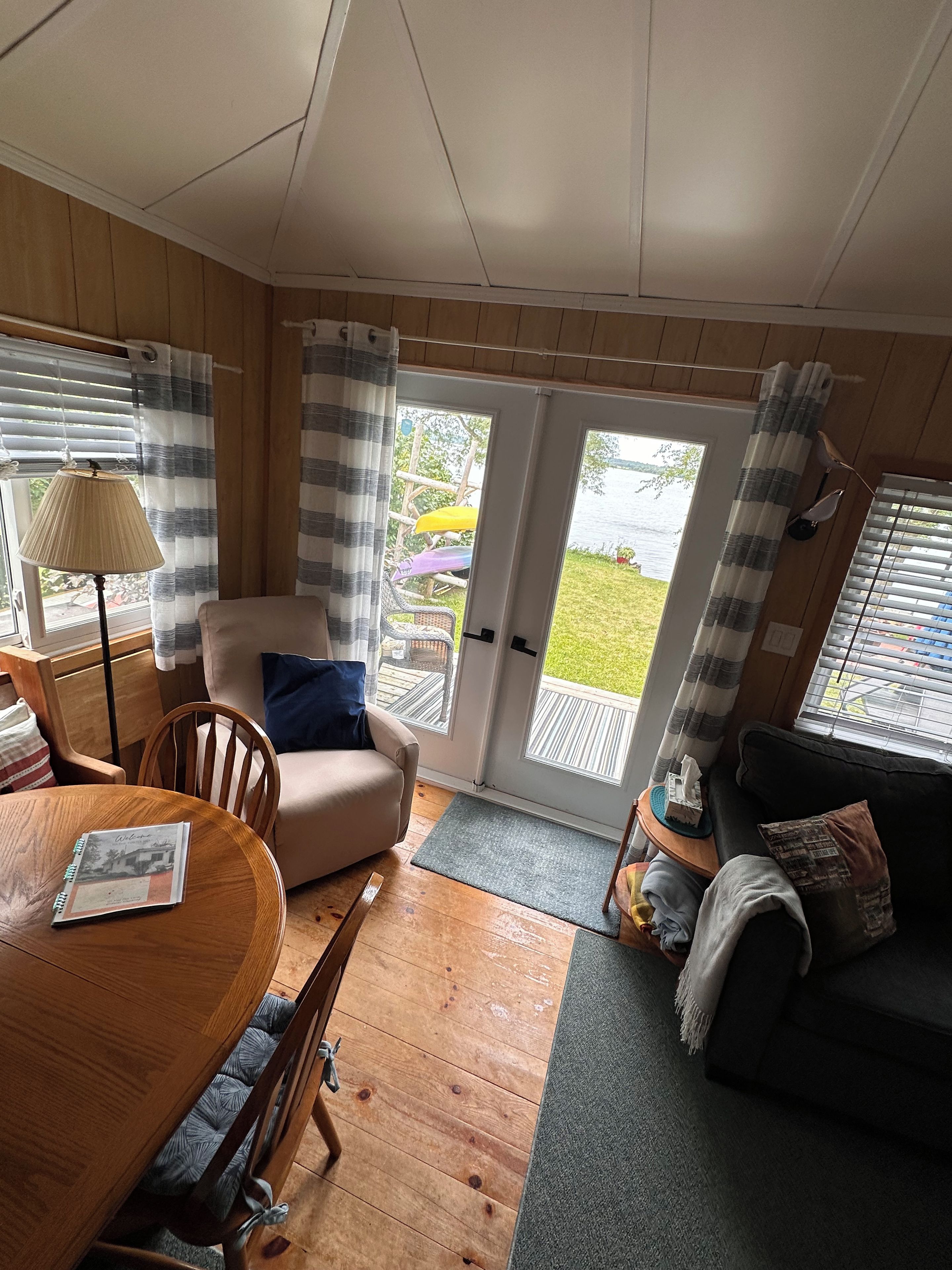 Une pièce chaleureuse avec un plancher en bois, une table ronde sur laquelle se trouve un carnet, un fauteuil beige, un canapé foncé et une vue sur un jardin, visible à travers une porte vitrée.