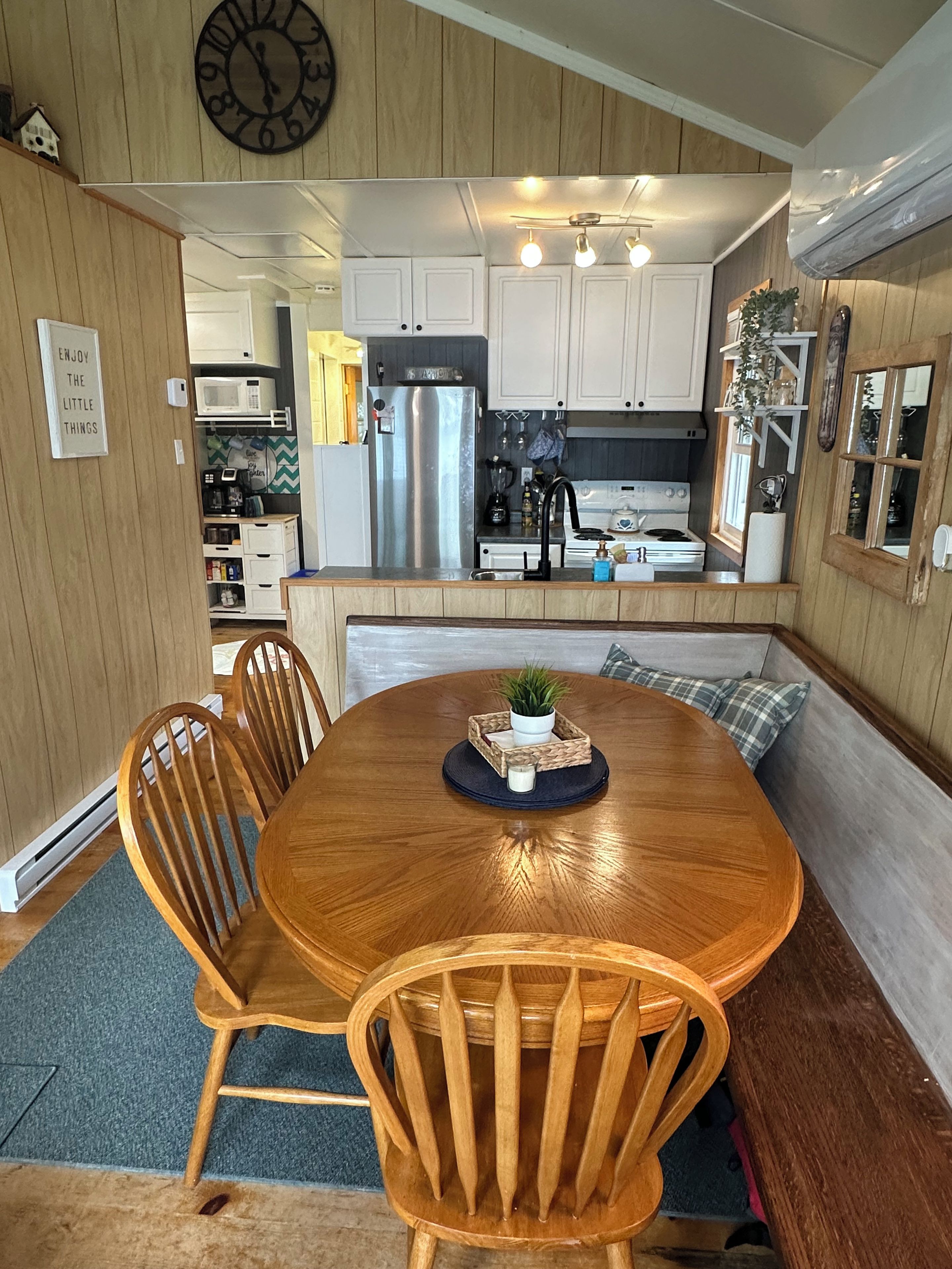 A dining area with a wooden table and chairs, a clock on the wall, and a kitchen in the background.