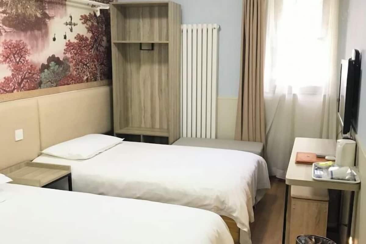A hotel room with two beds, a wooden wardrobe, a radiator, a desk with a chair, and a TV.