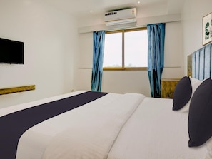 A hotel room with a bed, a mounted TV, an air conditioning unit, and a window with curtains.
