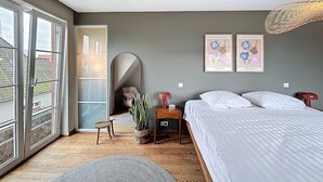 A bedroom with a large bed, a small table with a lamp, a potted plant, and a mirror. There are two framed pictures on the wall and a window with a view of buildings outside. - Villa Charme - Koksijde (Koksijde)